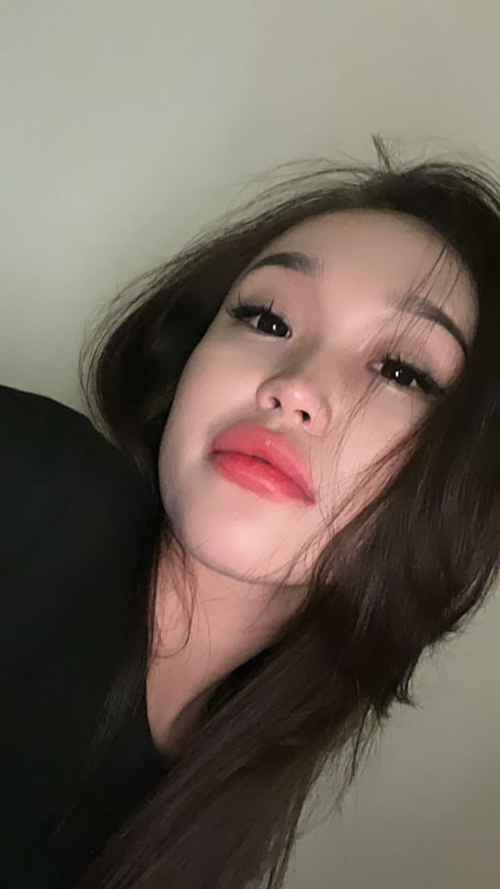 Close-up selfie of a woman with long dark hair and red lipstick, wearing a black top and tilting her head against a plain light-colored wall.