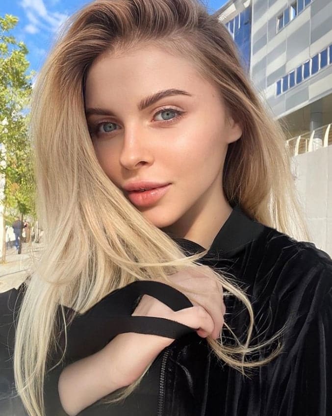 Eliza stands outdoors in a black velvet top with a shoulder cutout, positioned before a modern building and trees under a clear blue sky.