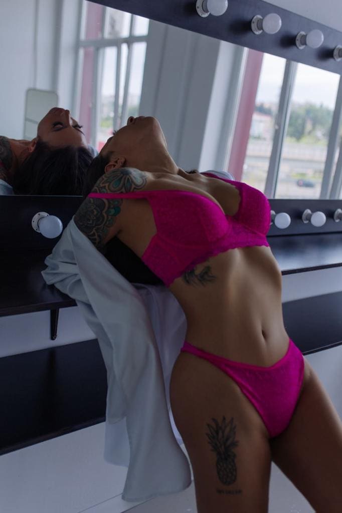 A woman leans back against a vanity mirror with light bulbs in bright pink lace lingerie, with a white shirt draped on the counter behind her.