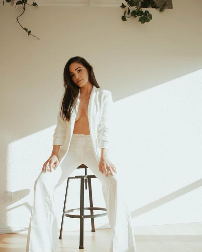 Erika sits on a black stool in an unbuttoned white blazer and trousers, exposing her midriff in a bright room with a wall plant.