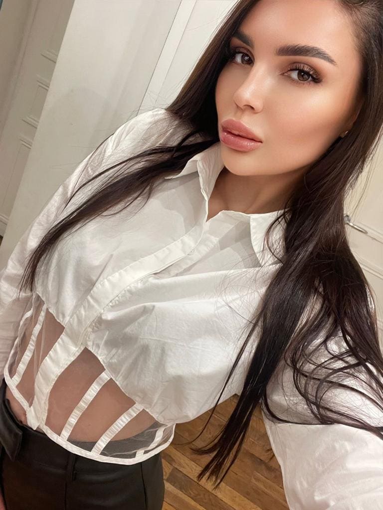 A woman with long dark hair poses in a white shirt with sheer side panels and dark trousers, standing on a wooden floor against white walls.