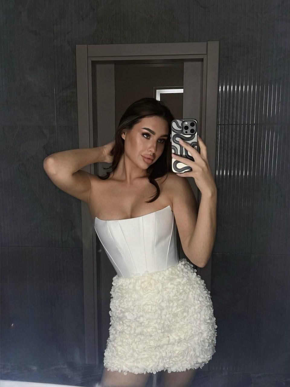 Mirror selfie in a white corset top and textured skirt against a dark paneled wall.