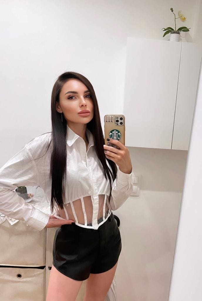 Zaritta takes a mirror selfie in a white room, wearing a white cropped shirt with sheer midriff panel and black shorts, holding a phone.