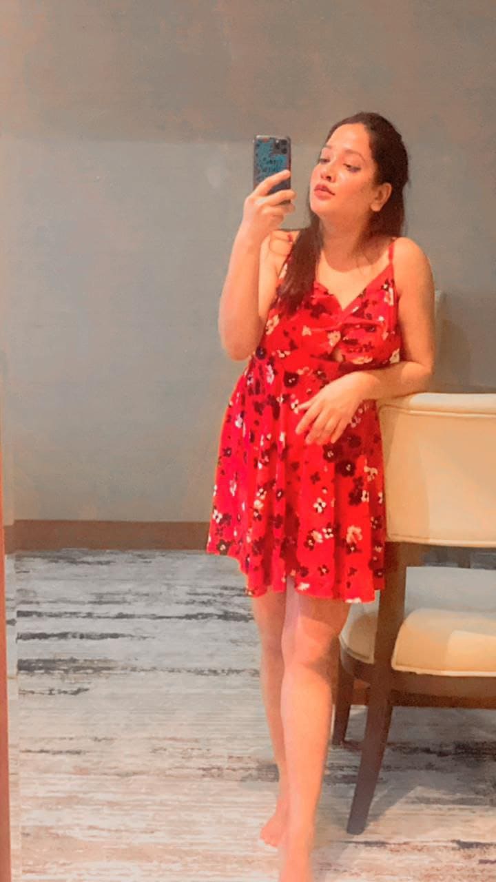 A barefoot woman in a short red floral dress leans against a chair, taking a mirror selfie in a room with a patterned rug.