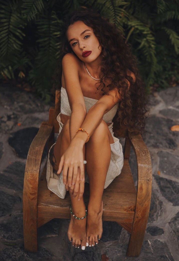 A woman sits on an outdoor wooden chair in a light strapless dress, arms crossed over knees, against green foliage and stone.