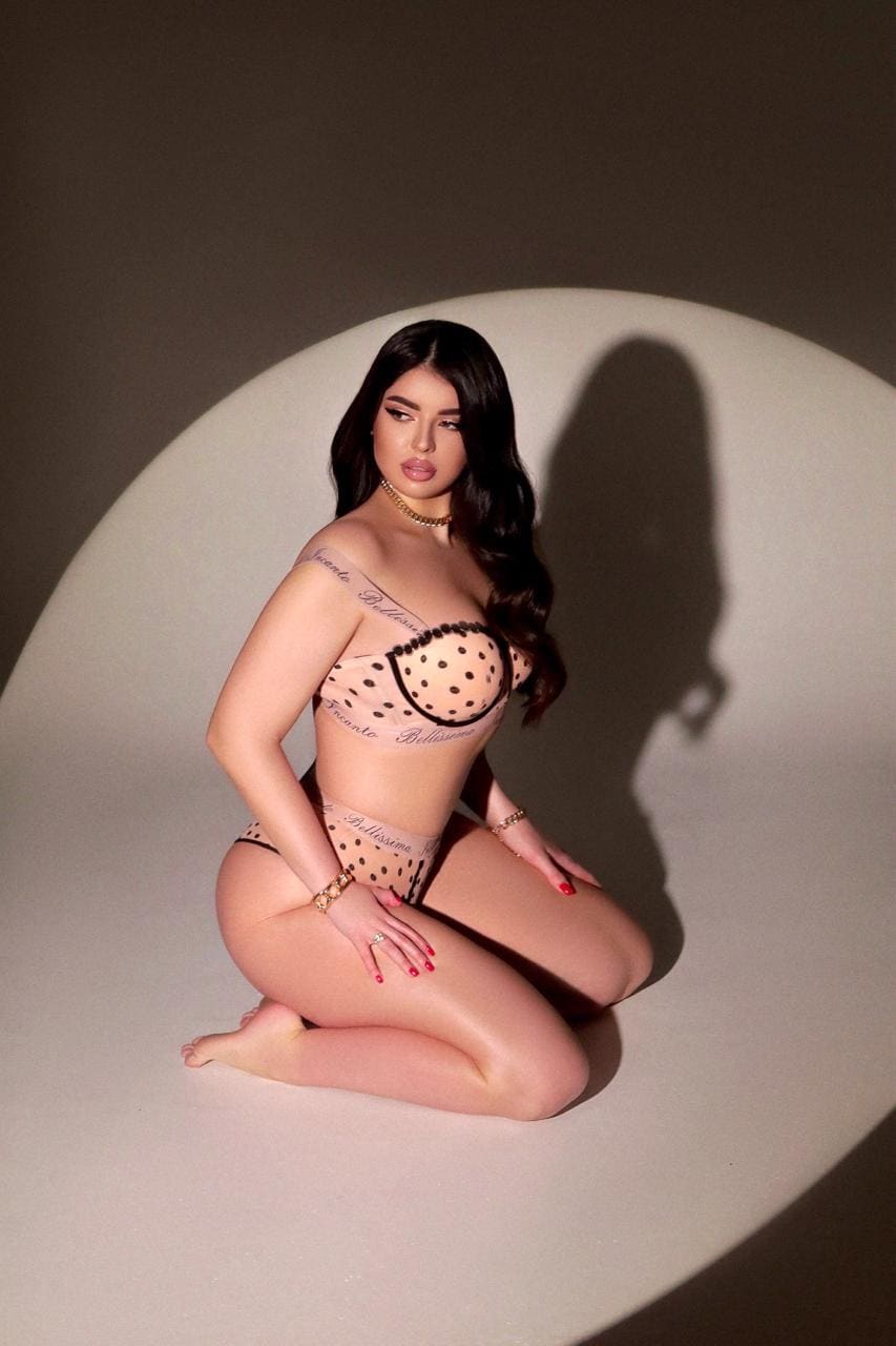 A woman kneels in a spotlight wearing a sheer polka-dot lingerie set, gold choker, and bracelets, with visible tattoos.