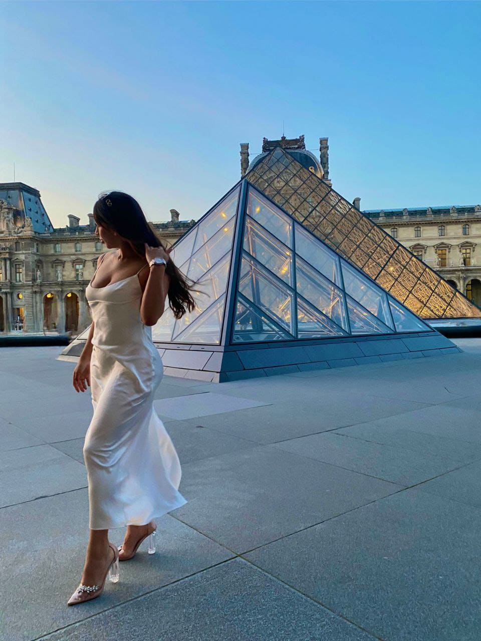 A woman in a white satin slip dress walks in profile on a stone plaza in front of a glass pyramid, touching her hair.