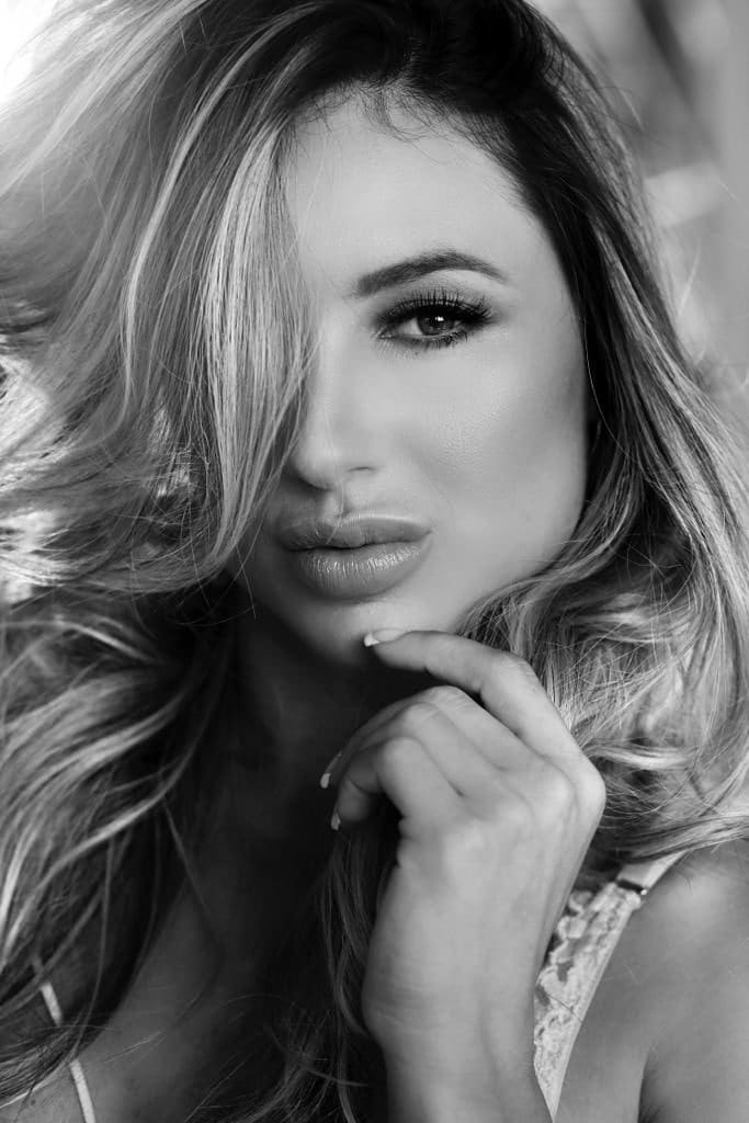 Black-and-white close-up portrait of a woman in a lace-trimmed top, hand near chin, against a blurred background.