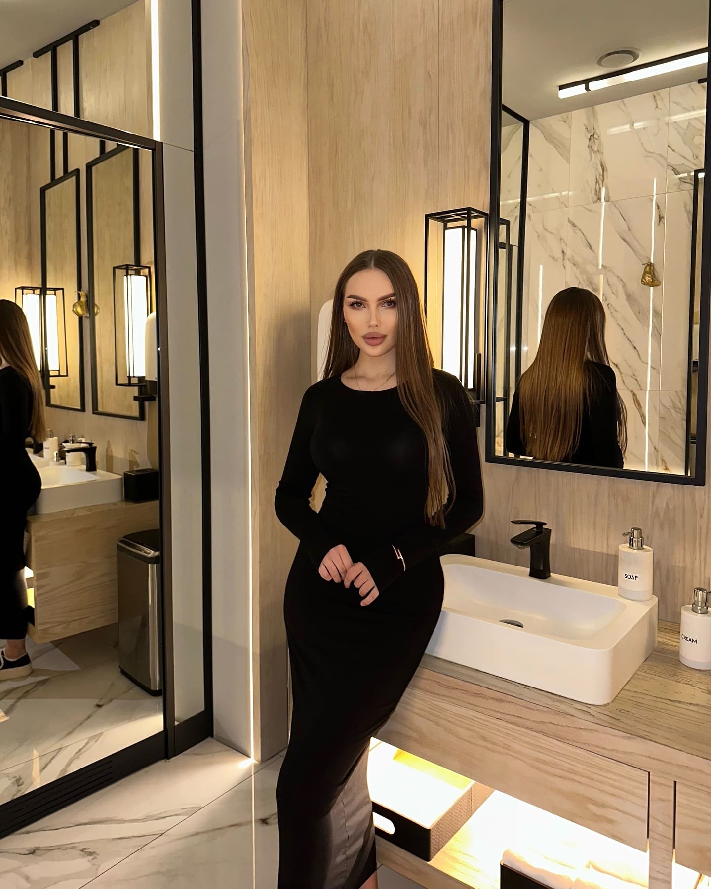 Lorita stands in a marble bathroom wearing a long-sleeved black dress, hands clasped in front of her.
