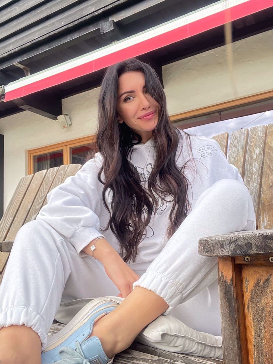 A woman sits on a wooden bench wearing a white hoodie, cuffed sweatpants, and light blue sneakers, with one leg crossed, in front of a building.