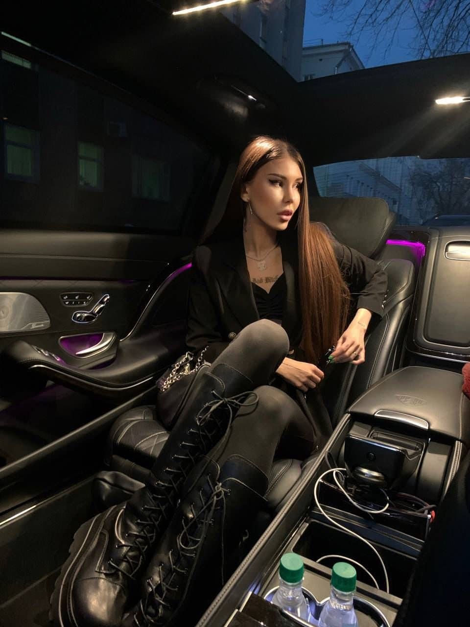 A woman sits in a car at night wearing a black blazer, leggings, and tall lace-up boots, with a chain-strap bag on her lap.