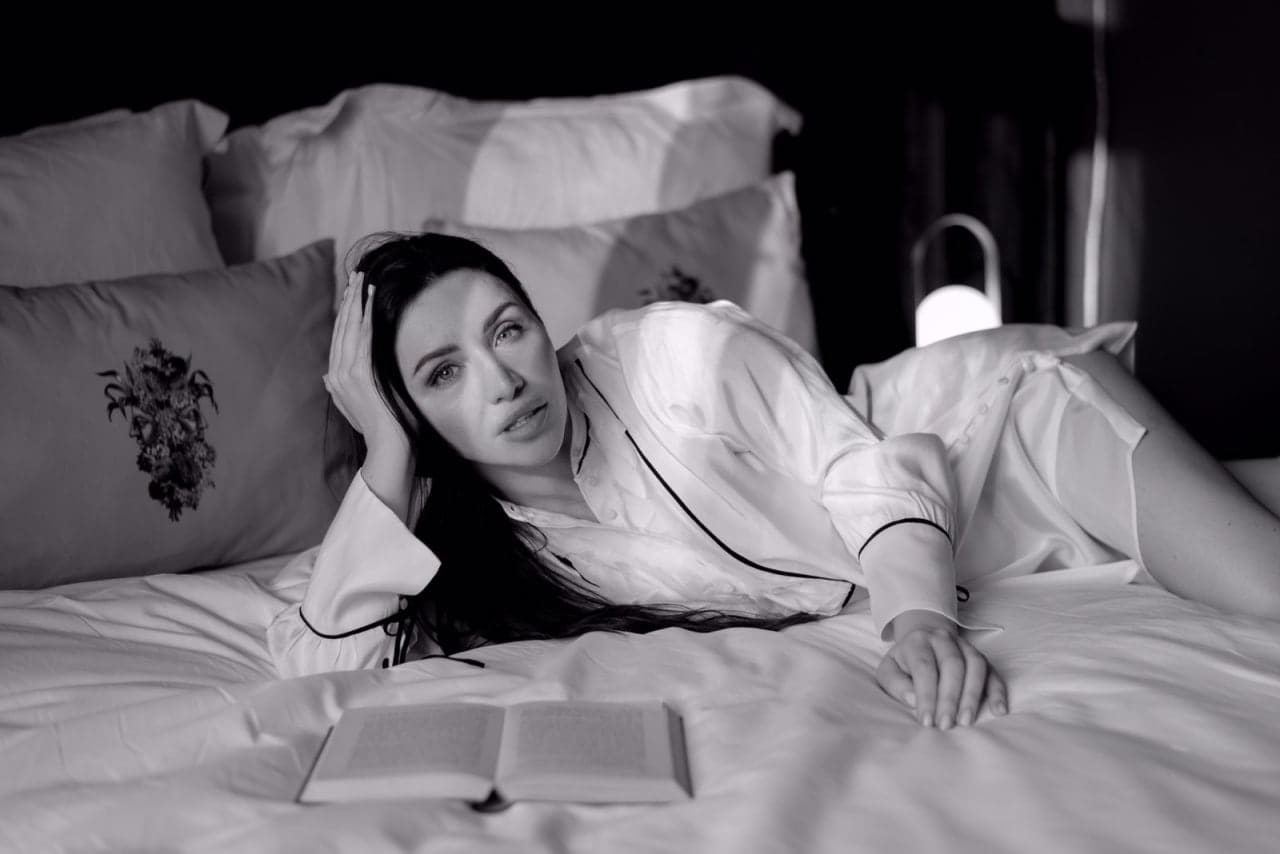 A woman lies on her side on a bed with white linens, wearing a white button-up shirt with dark piping, one hand on her head, an open book nearby.