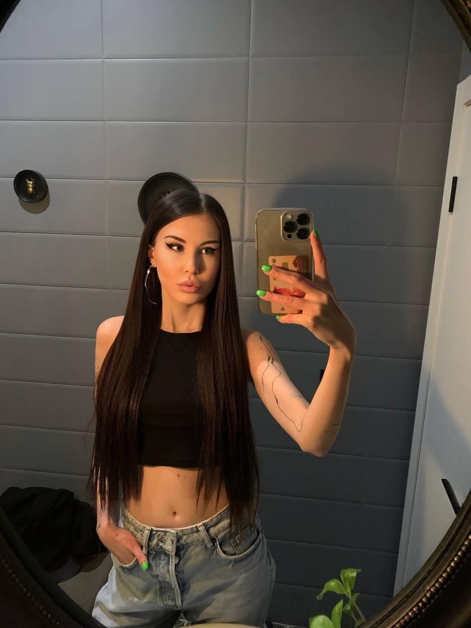Amanda takes a mirror selfie in a tiled room, wearing a black cropped tank top and light-wash jeans with her midriff exposed.