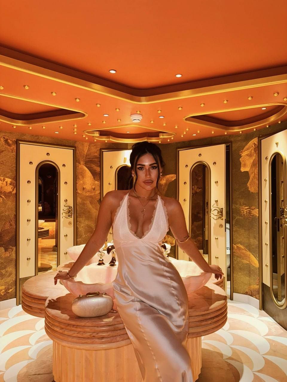 Woman in a satin slip dress sits on a wooden table in a room with orange walls.