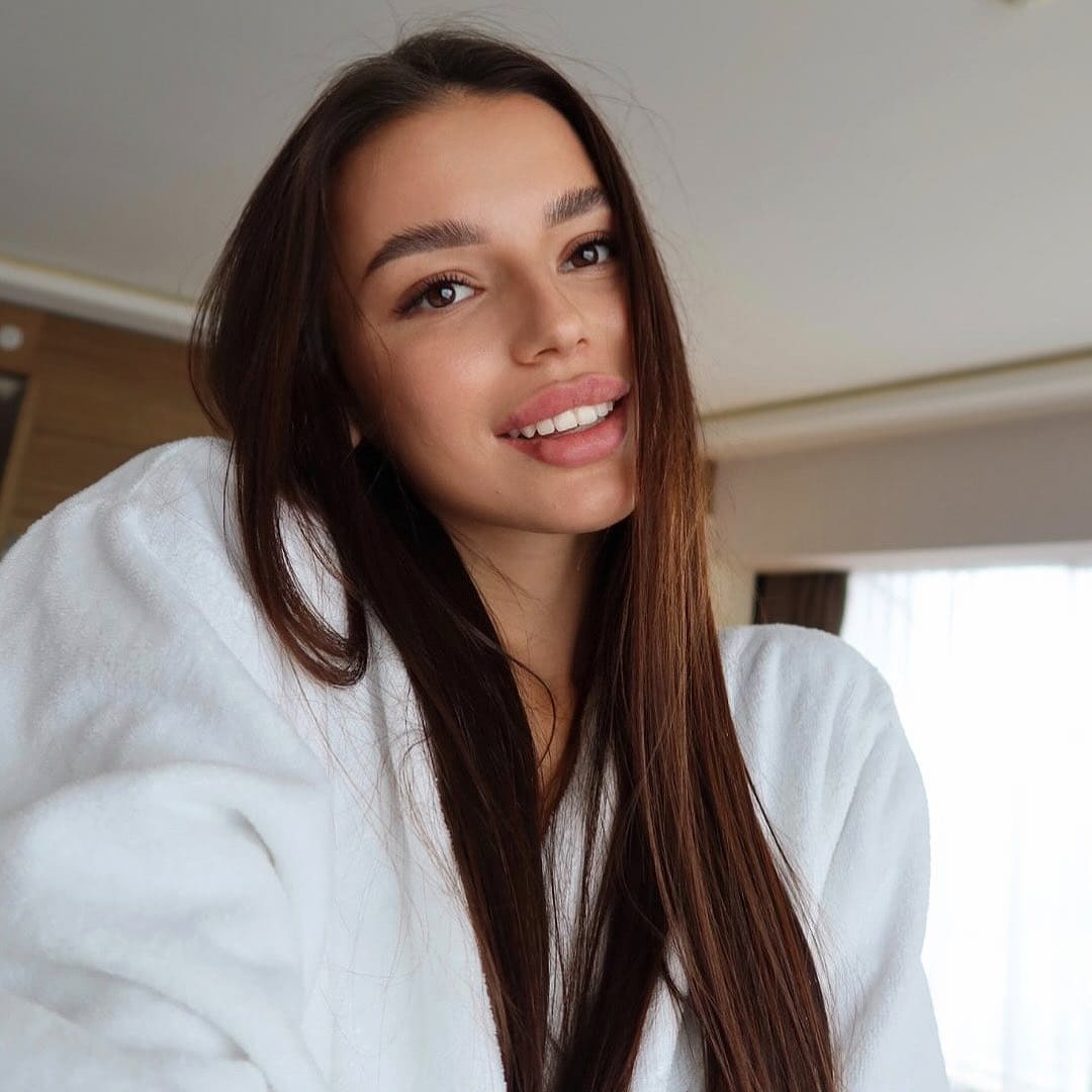 Elen smiles at the camera wearing a white fluffy robe in an indoor room with a window behind her.