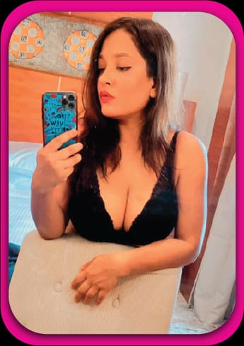 A woman sits on a bed taking a mirror selfie in a black lace bralette, with a quilted wall hanging behind her.