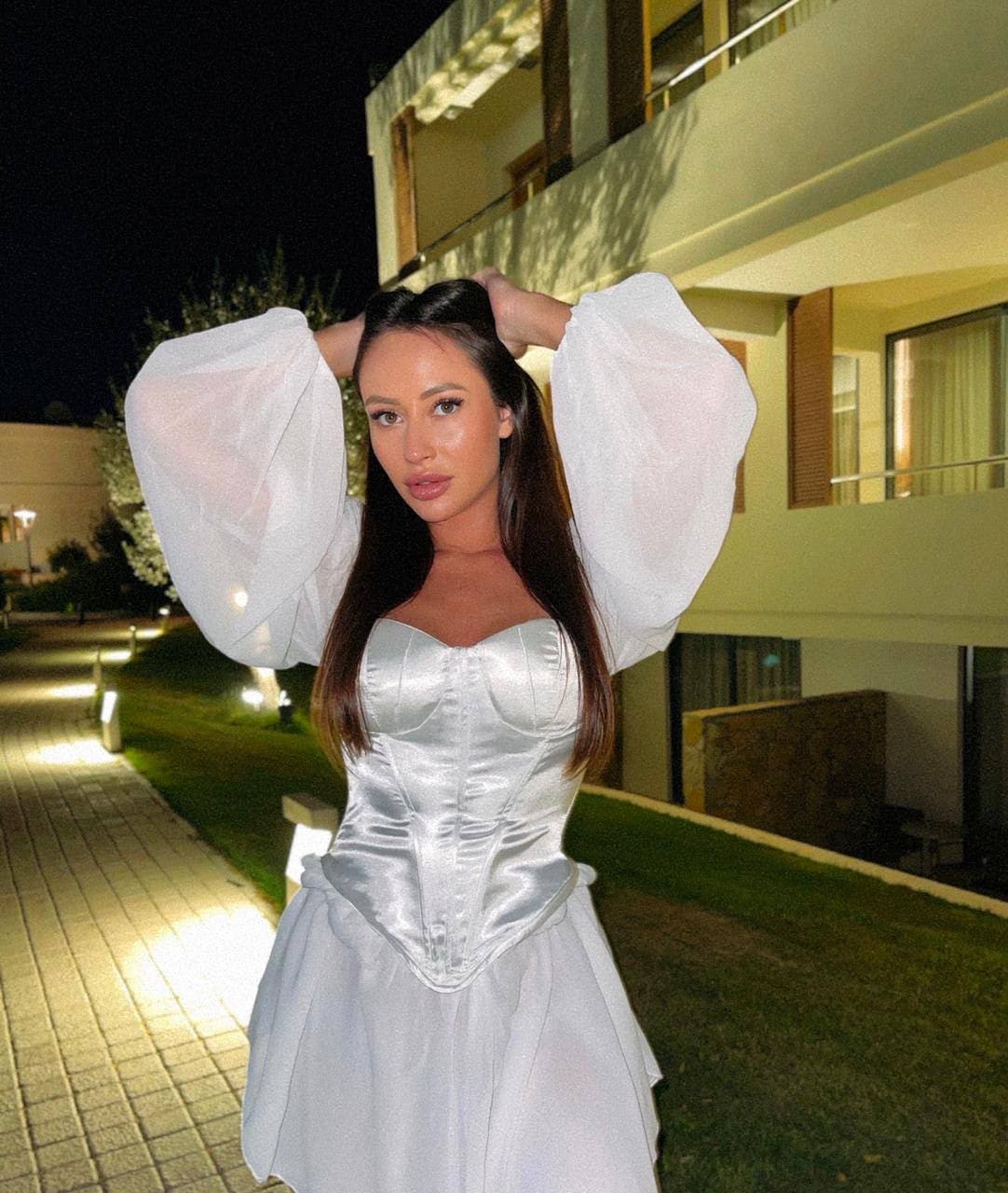 A woman stands outdoors at night in a white satin corset and skirt with sheer puff sleeves, hands raised behind her head.