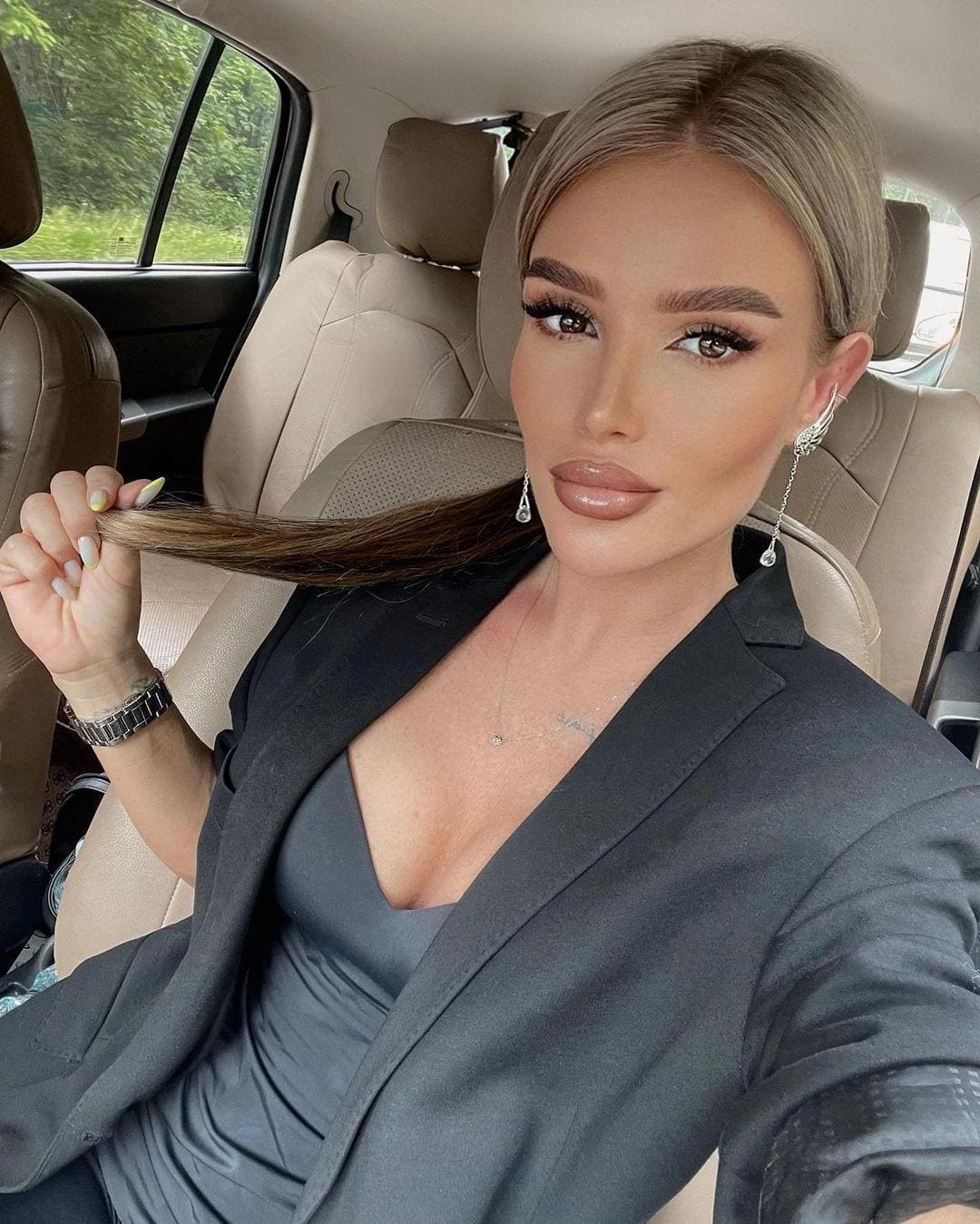A woman in a dark grey blazer and low-cut top sits in a car back seat, holding a strand of blonde hair while looking at the camera.