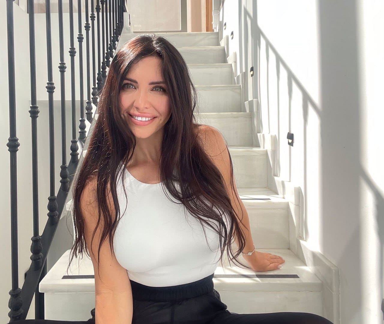 Ani sits on a white marble staircase with black railings, wearing a white sleeveless top and black pants, smiling in bright natural light.