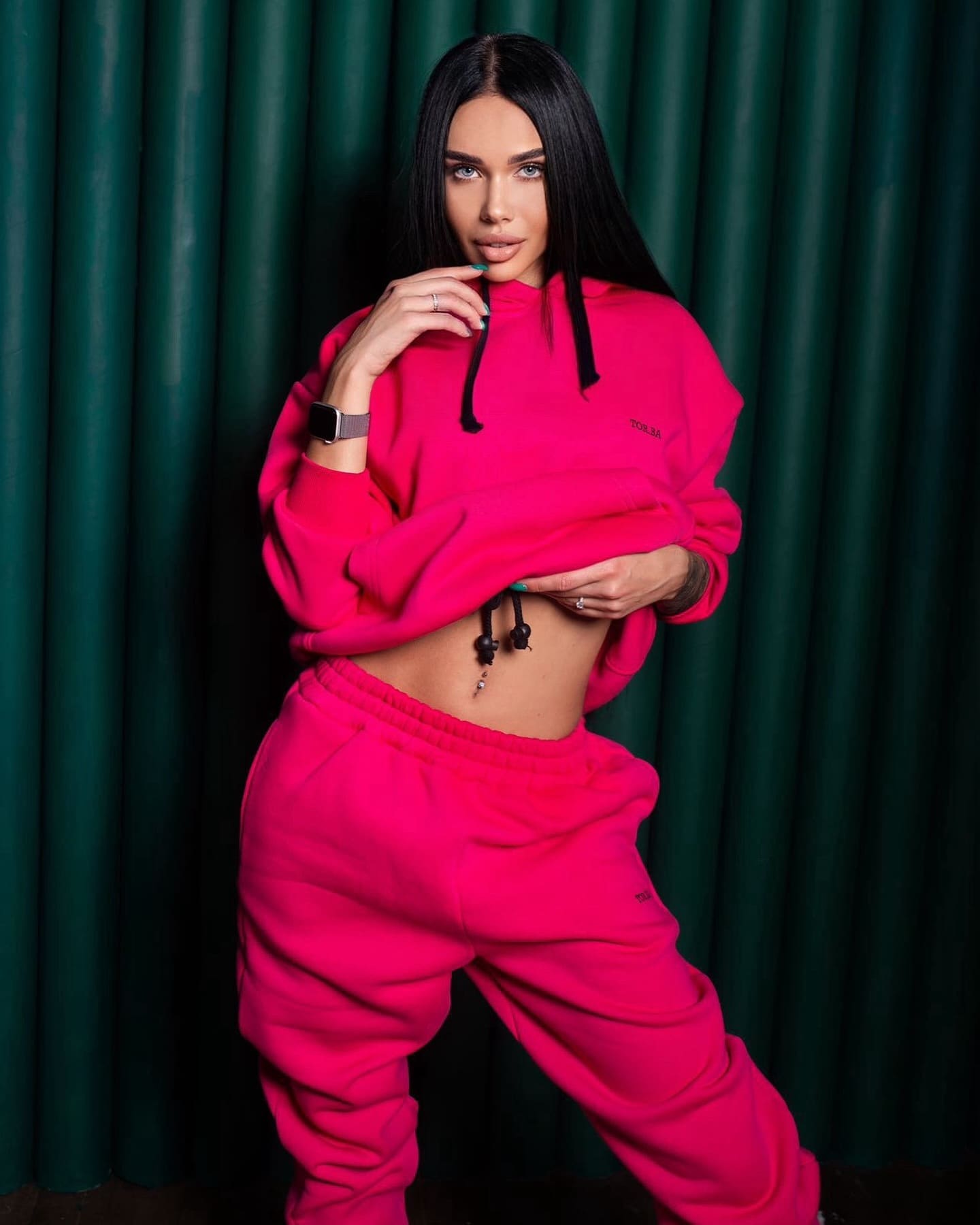 Taya stands before a dark green curtain in a pink cropped hoodie and sweatpants, one hand near her chin and the other on her waist.