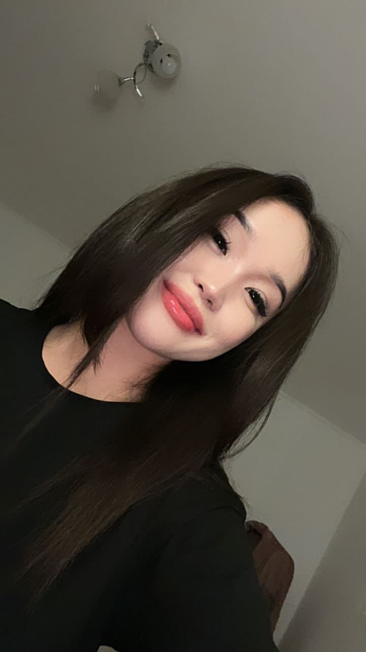 A woman with long dark hair and red lipstick tilts her head while wearing a black top in a room with a white ceiling light fixture above.