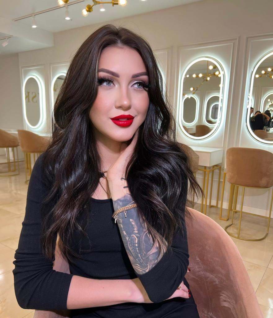 Dana sits in a salon chair wearing a black long-sleeved top, her hand near her neck, surrounded by oval mirrors and gold stools.