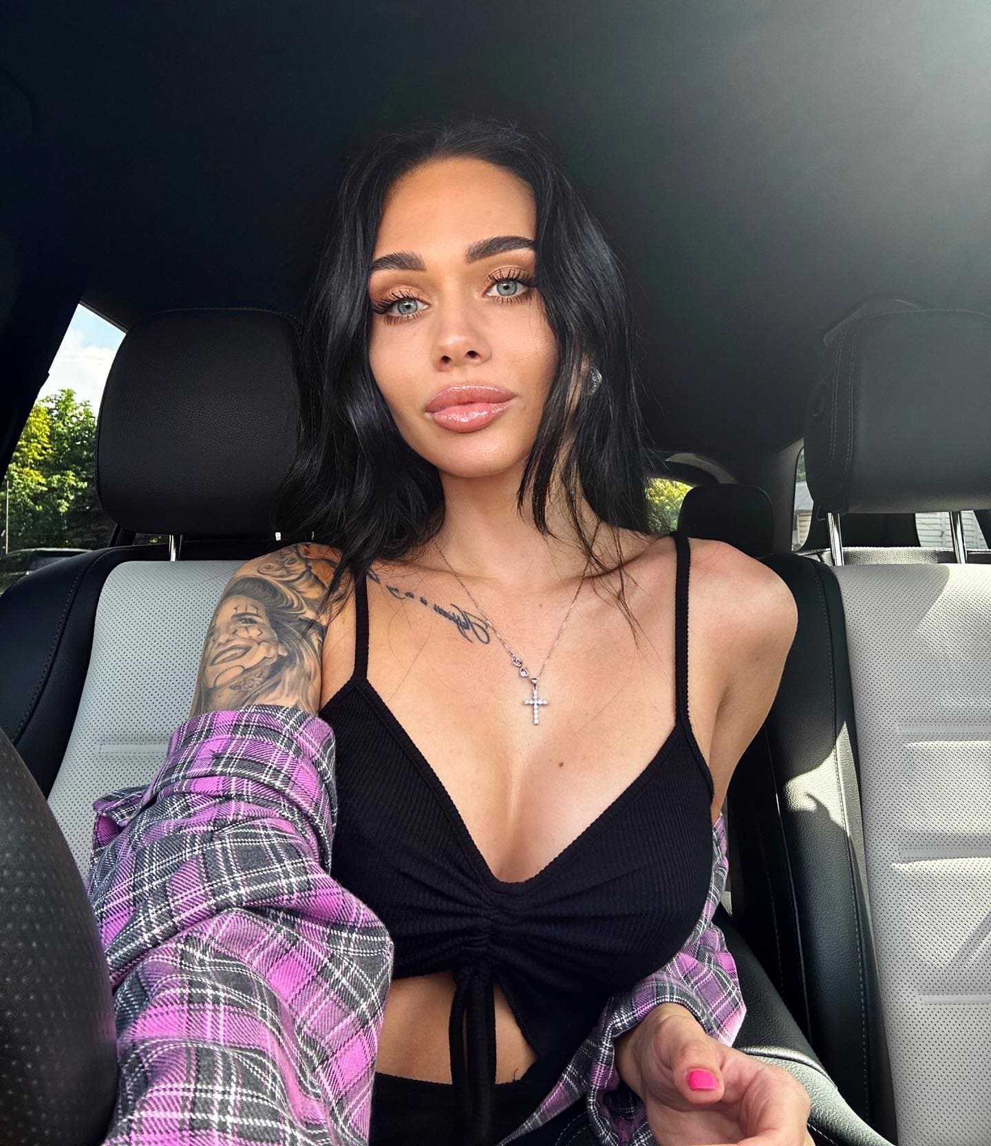 A woman sits in a car back seat wearing a black crop top, plaid shirt on arms, cross necklace, and a shoulder tattoo.