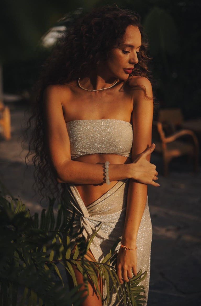 A woman stands in warm light wearing a sparkly silver two-piece outfit and beaded jewelry, arms crossed, with ferns in the foreground.
