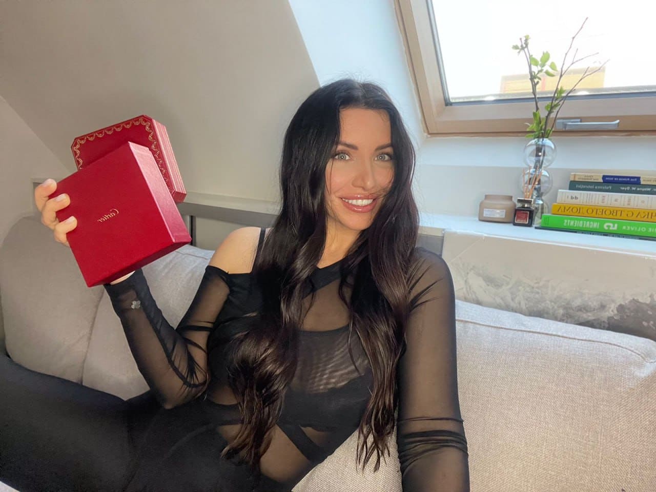 A woman sits on a light couch near a window, wearing a sheer black top with cut-outs, holding a red box in one hand.