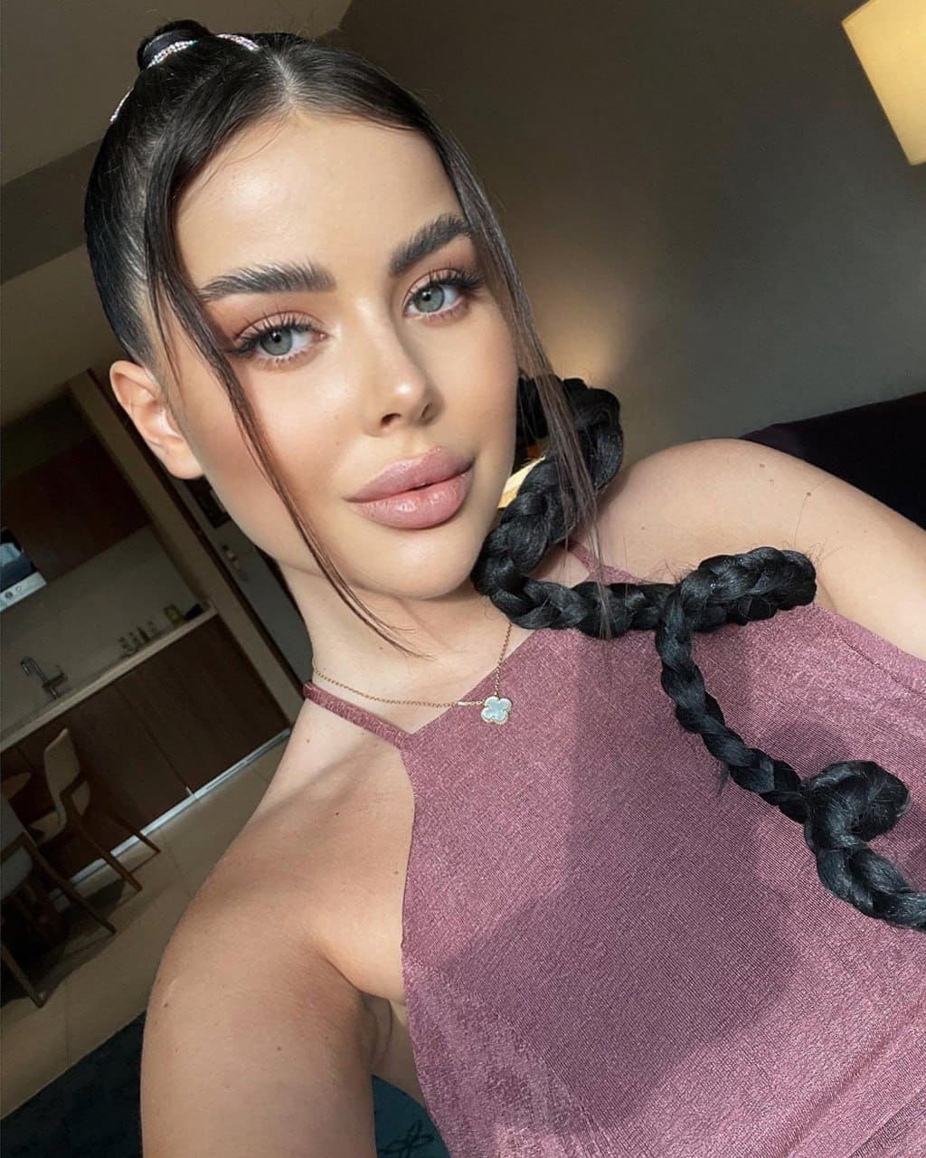 Woman with a high bun and long braid wears a sleeveless mauve top and thin necklace, posing indoors near a kitchen area.