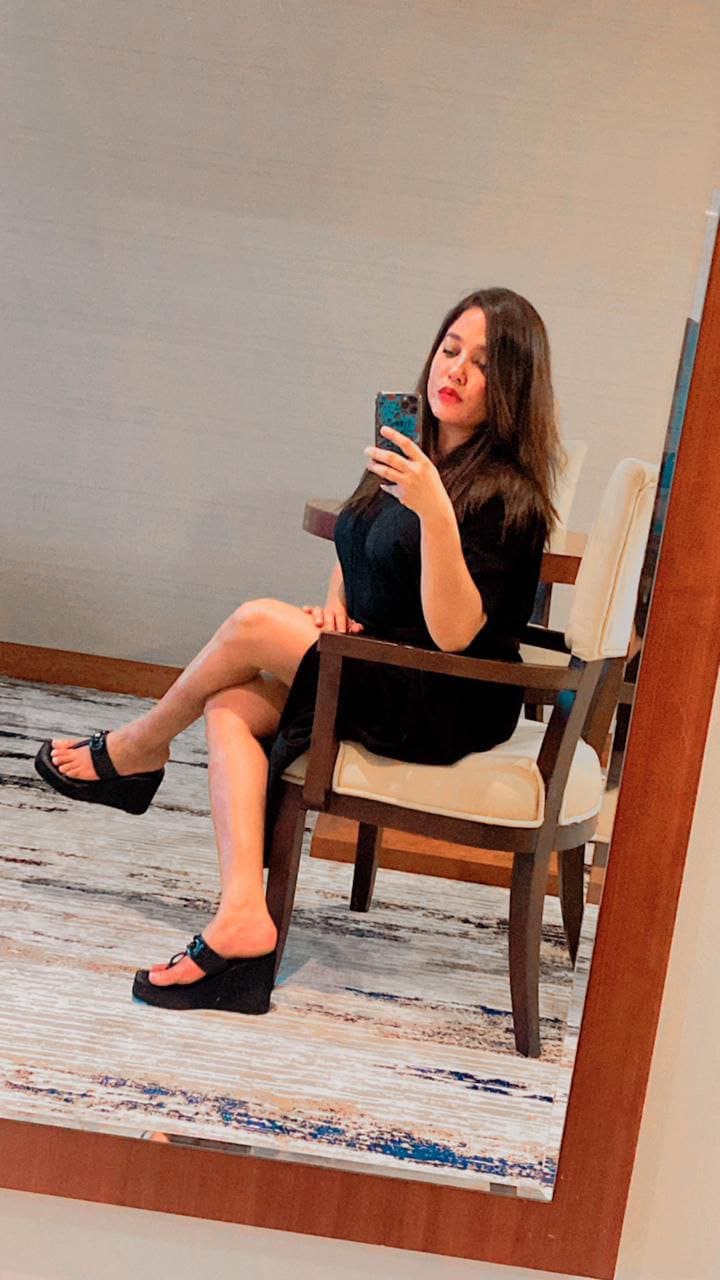 A woman sits on a wooden chair with a cream cushion, wearing a black dress and platform sandals, looking at her smartphone.