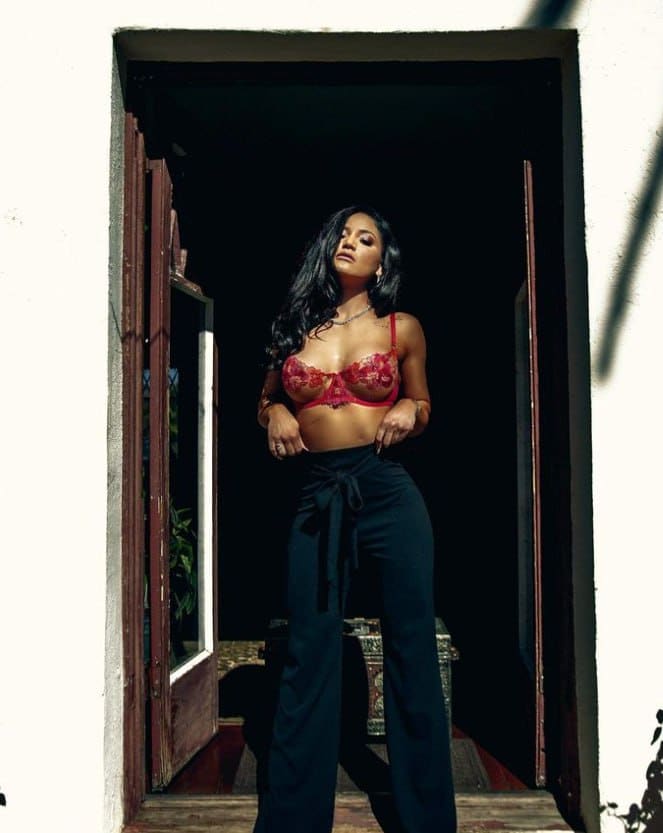 A woman stands in a doorway wearing a red lace bra and dark high-waisted trousers, with one hand resting on her hip.