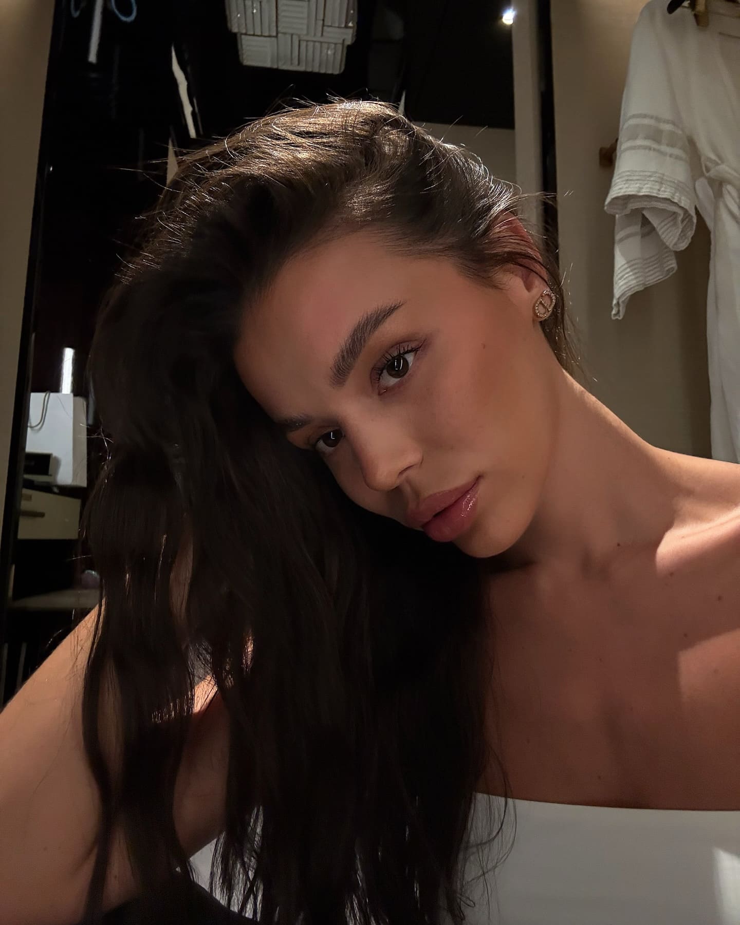A woman with long dark wavy hair tilts her head, wearing a white strapless top and stud earring near a mirror.