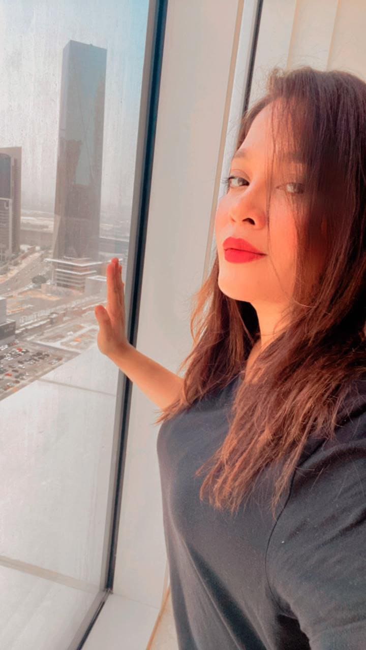 ANAM stands by a window overlooking a city skyline, wearing a dark grey top with one hand on the glass.