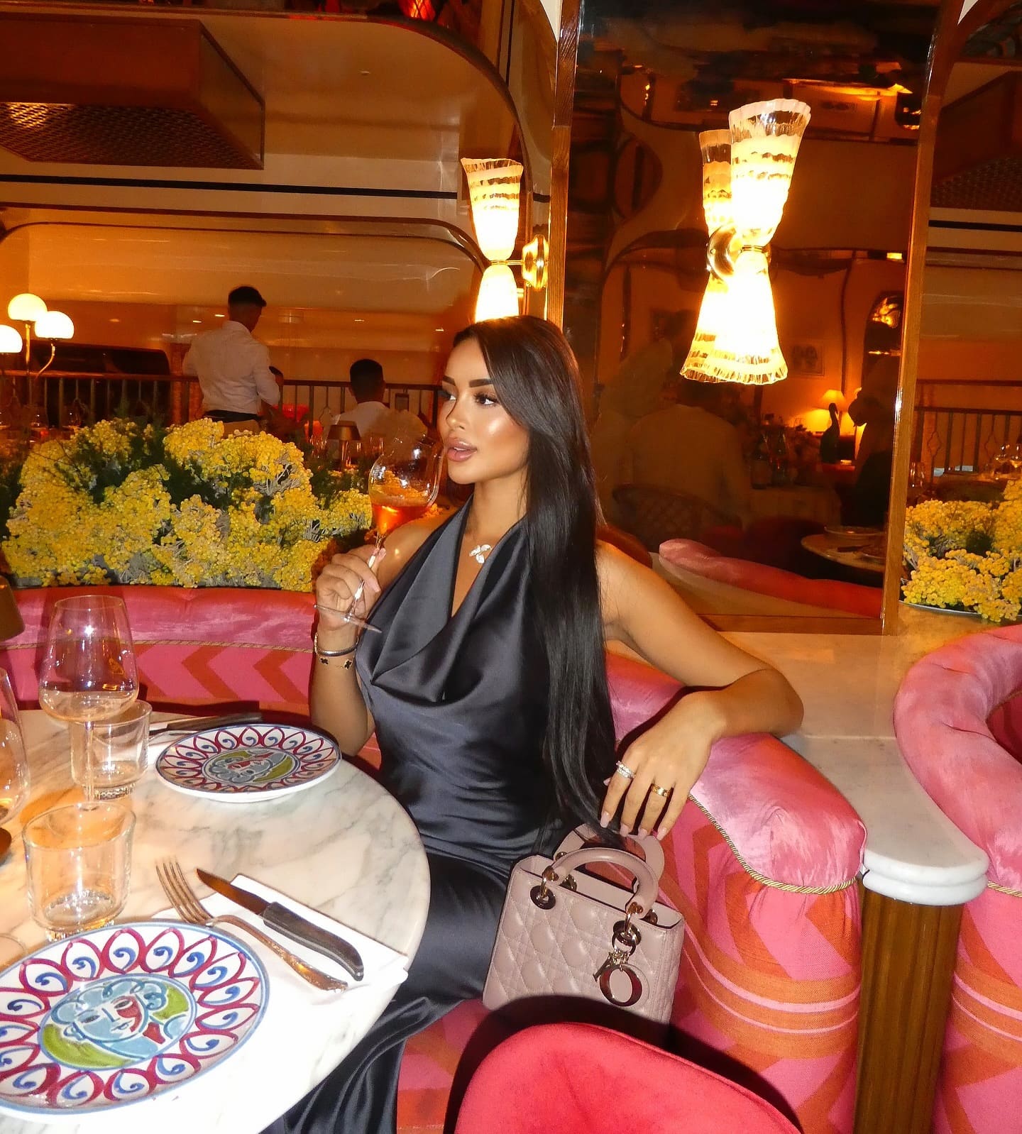 Seated at a marble restaurant table, a woman in a black satin dress holds a glass of rosé wine, with a quilted handbag on her lap.