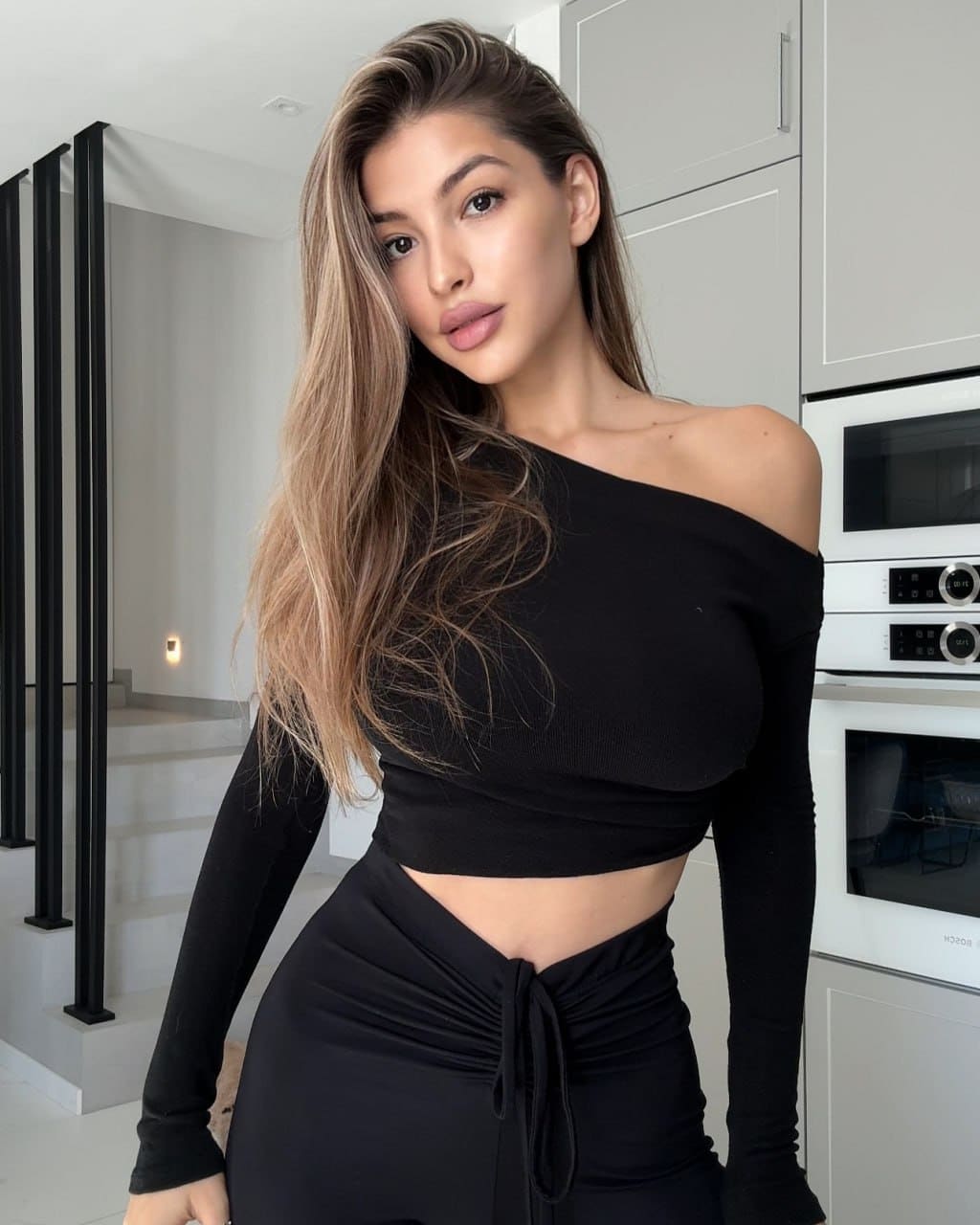 Lia stands in a modern kitchen wearing a black one-shoulder crop top and high-waisted black skirt with a front tie.