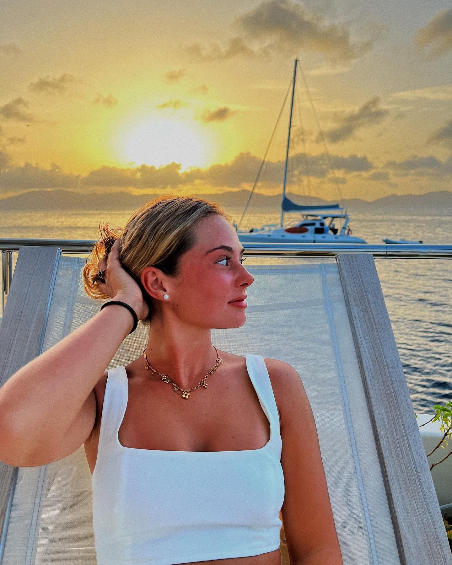 A woman in a white crop top and gold necklace stands on a boat deck at sunset, hand raised to her hair, with a sailboat in the background.