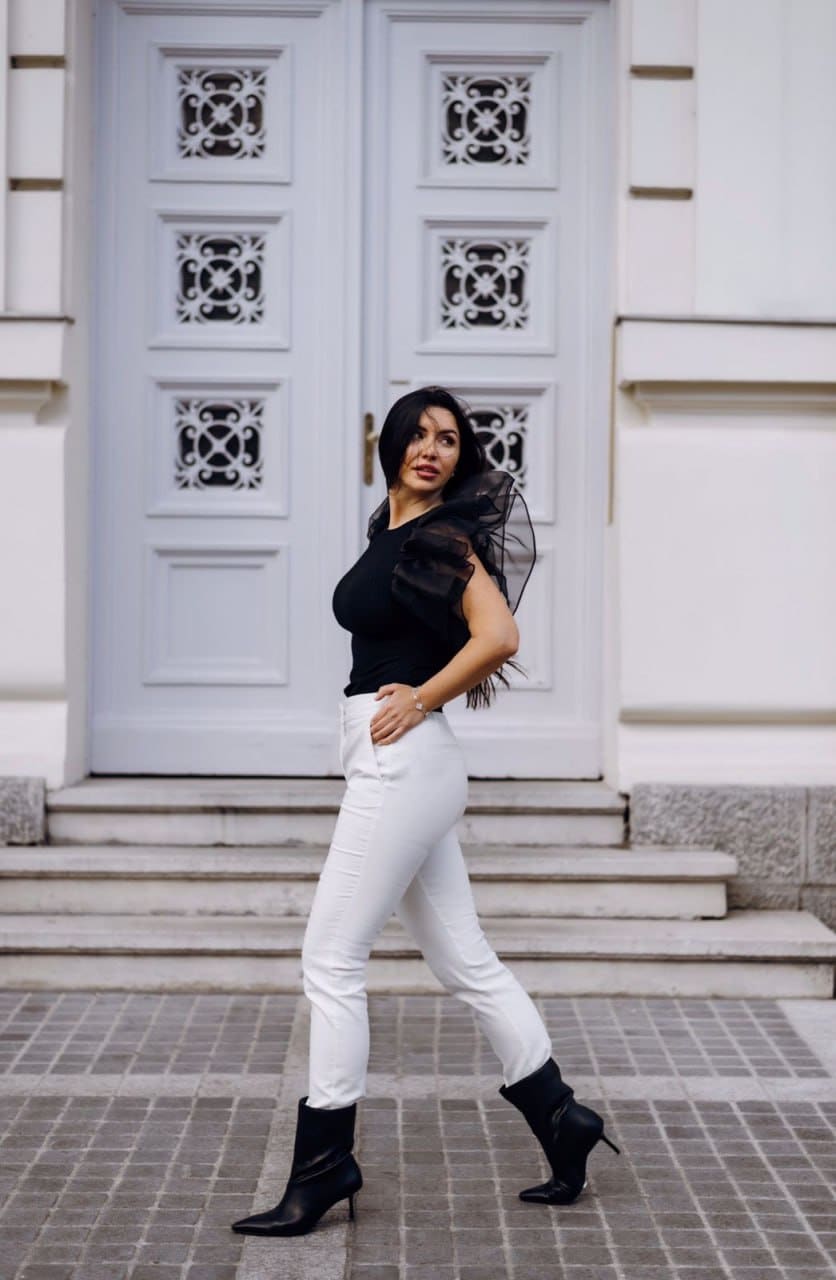 A woman stands in a three-quarter pose on pavement before a white door, wearing a black top with sheer sleeves, white trousers, and black ankle boots.