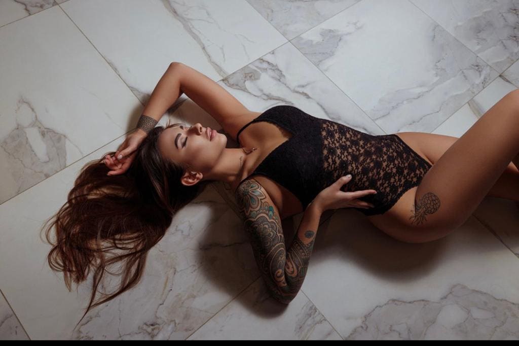 A woman lies on her side on a white marble-tiled floor in a black lace bodysuit, one arm raised behind her head, showing tattoos on her arm and thigh.