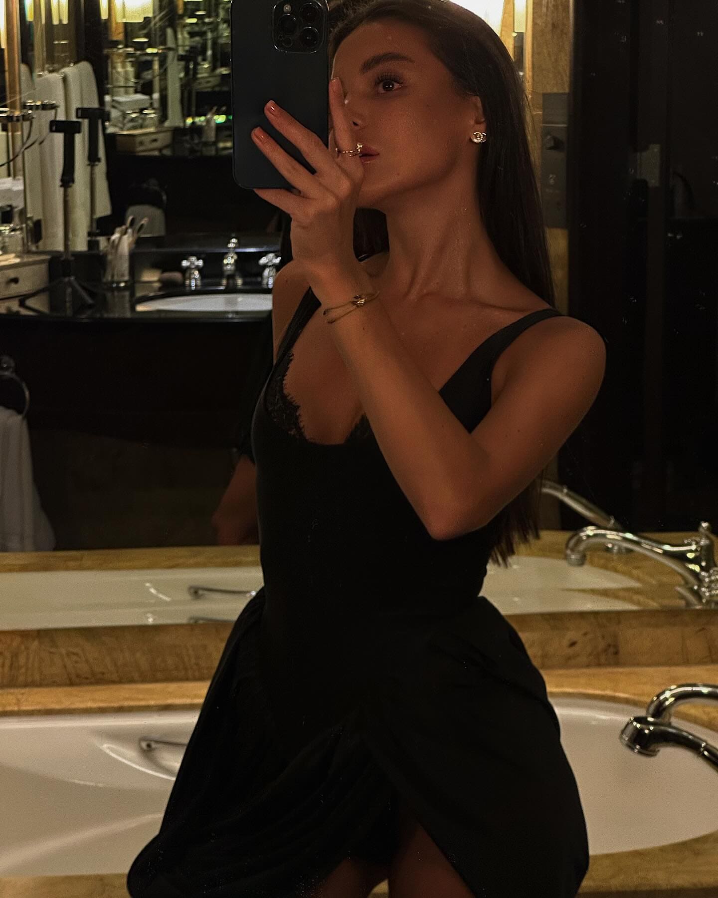 A woman takes a mirror selfie in a black lace dress beside a white bathtub, holding a phone to her face.