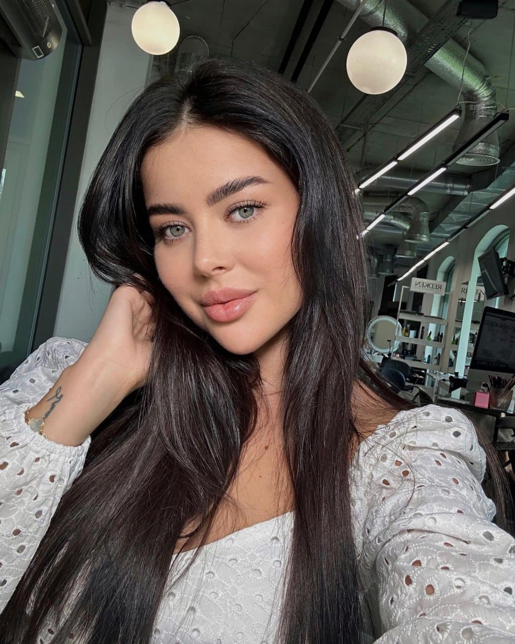 Virmi in a white eyelet blouse with a square neckline, hand raised to her hair, in a bright indoor space with exposed pipes and pendant lights.