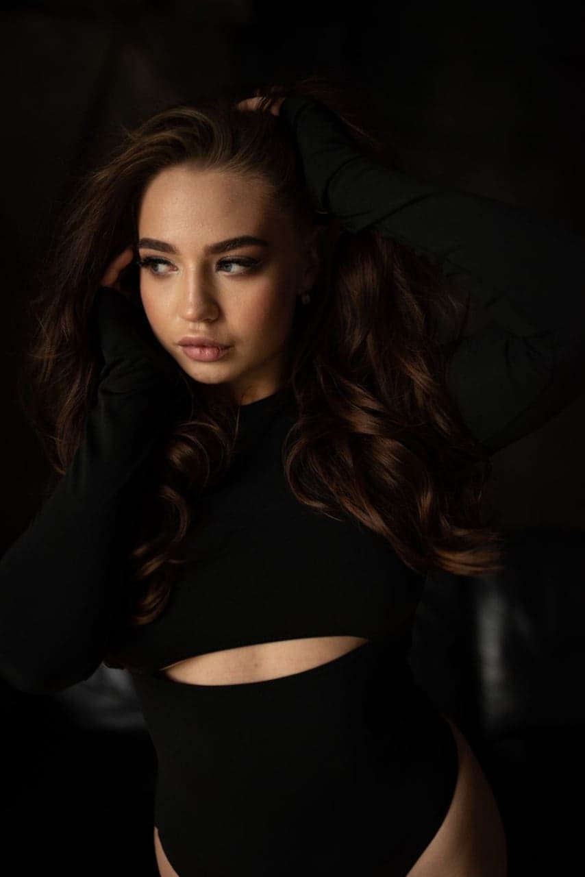 Emma poses with hands raised to her head, wearing a black long-sleeved bodysuit with a midriff cutout against a dark background.