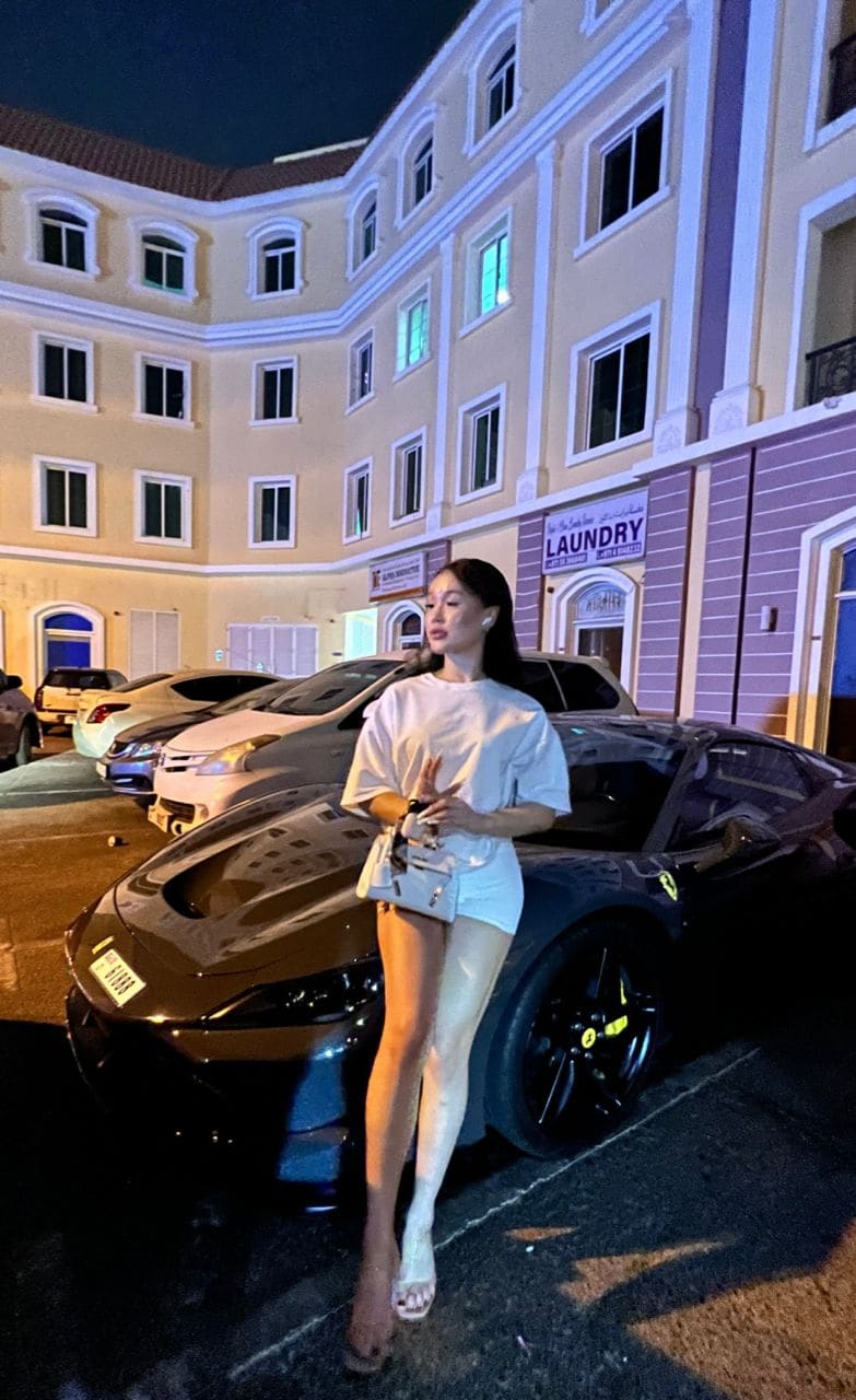 A woman in a loose white top, white high-heeled sandals, and a crossbody bag leans against a dark sports car in a parking lot at night.