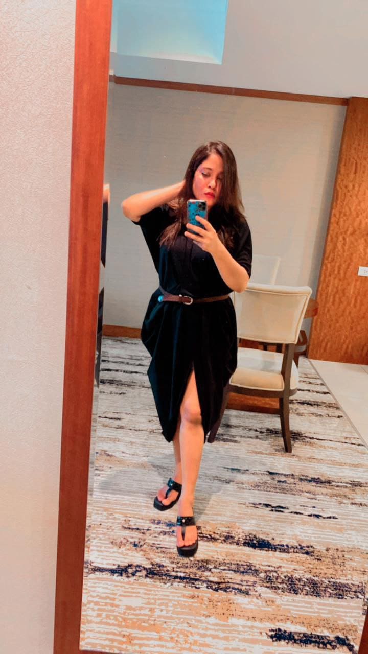 A woman takes a mirror selfie in a black dress with a high slit and brown belt, wearing platform sandals in a room with a patterned rug.