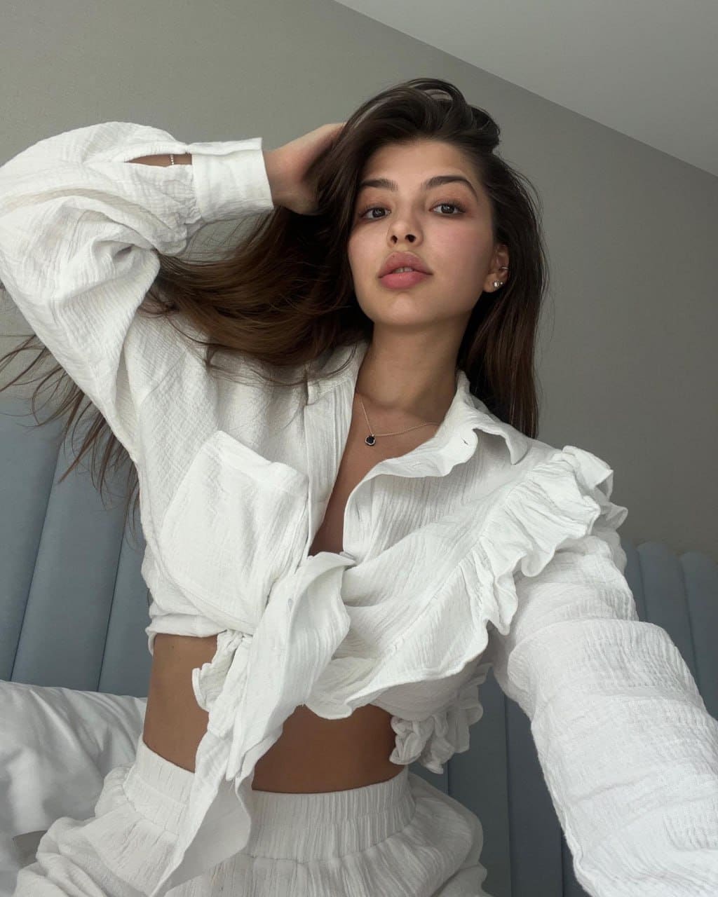 A woman sits against a gray headboard in a white ruffled blouse and matching pants, with one hand raised to her hair.