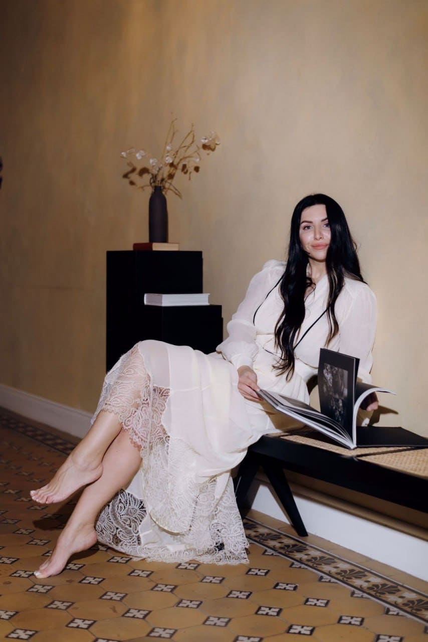 A woman sits barefoot on a low bench in a white lace dress, holding an open book, in a room with a patterned carpet and dried flowers on a pedestal.