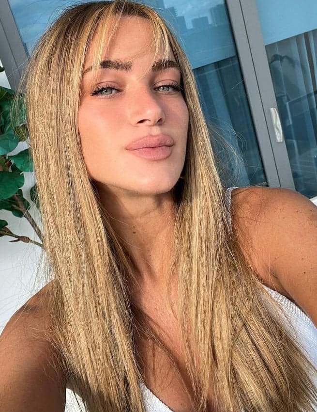 A close-up selfie of a woman in a white sleeveless top with a window and plant in the background.