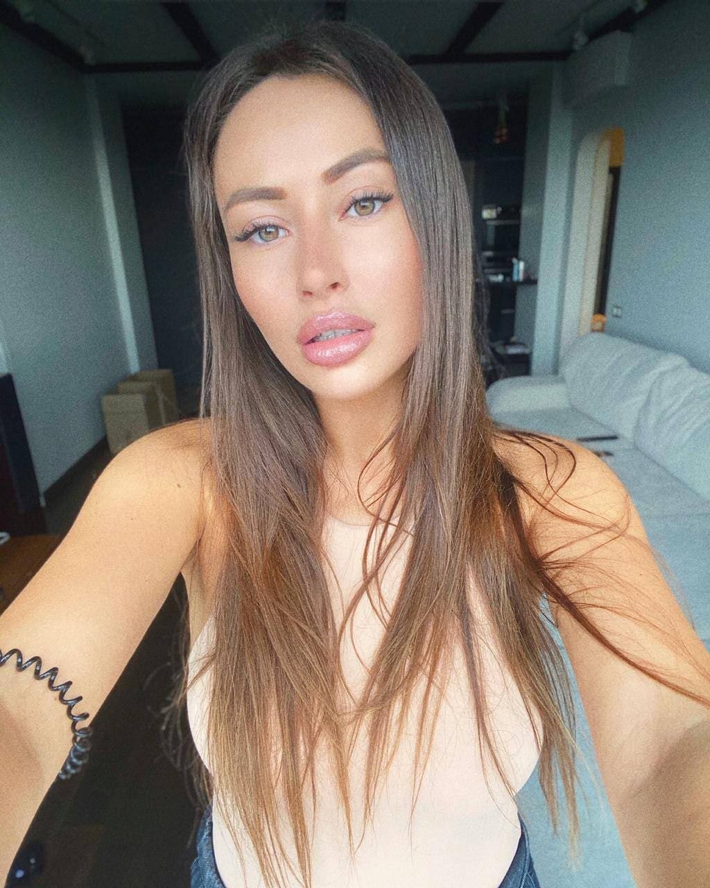 Kseniya takes a selfie in a living room, wearing a light top with a deep neckline, with a sofa and kitchen visible behind her.