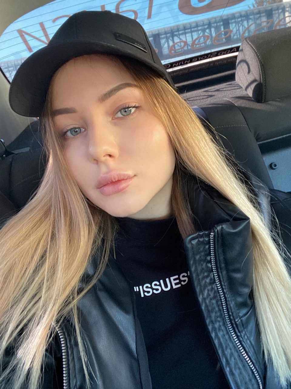 A woman wearing a black cap, leather jacket, and graphic tee sits in a car passenger seat.