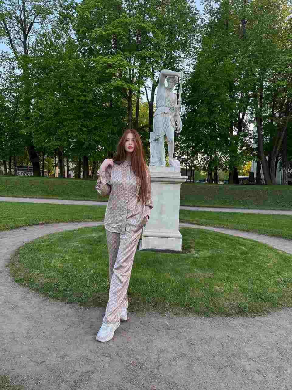 A woman with reddish-brown hair stands on a gravel path in a park, wearing a light patterned shirt and trousers with white sneakers near a statue.